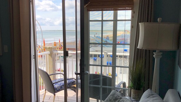 A room with a sliding glass door leading to a balcony with a view of the beach and a Ferris wheel. There is a sofa with patterned cushions and a lamp on a side table.