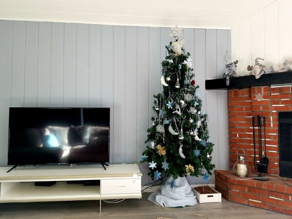 Een woonkamer met een tv op een witte dressoir, een kerstboom met decoraties, en een open haard van baksteen.