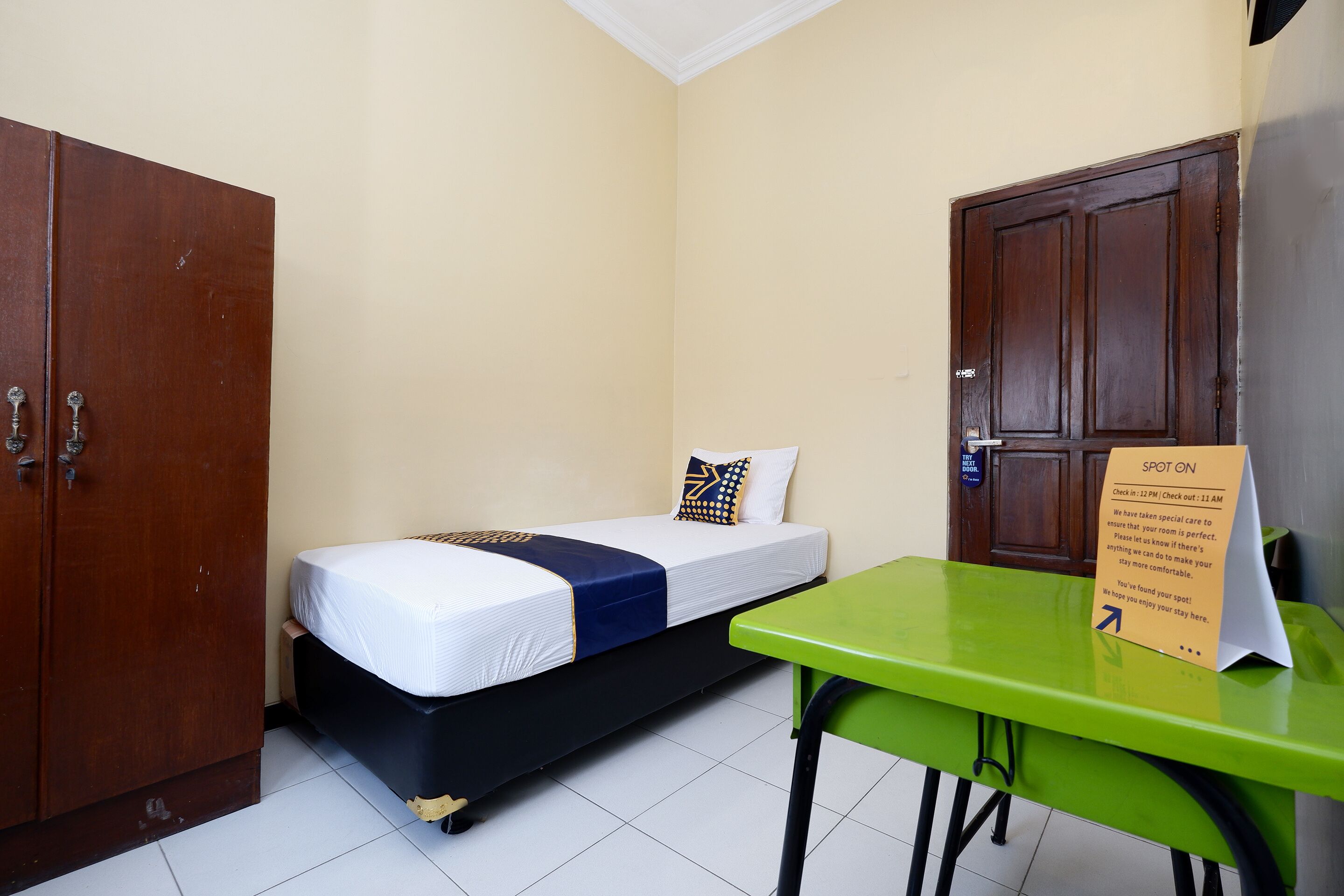 A small, single-bed room with a wooden wardrobe, a green table, and a sign indicating check-in times.