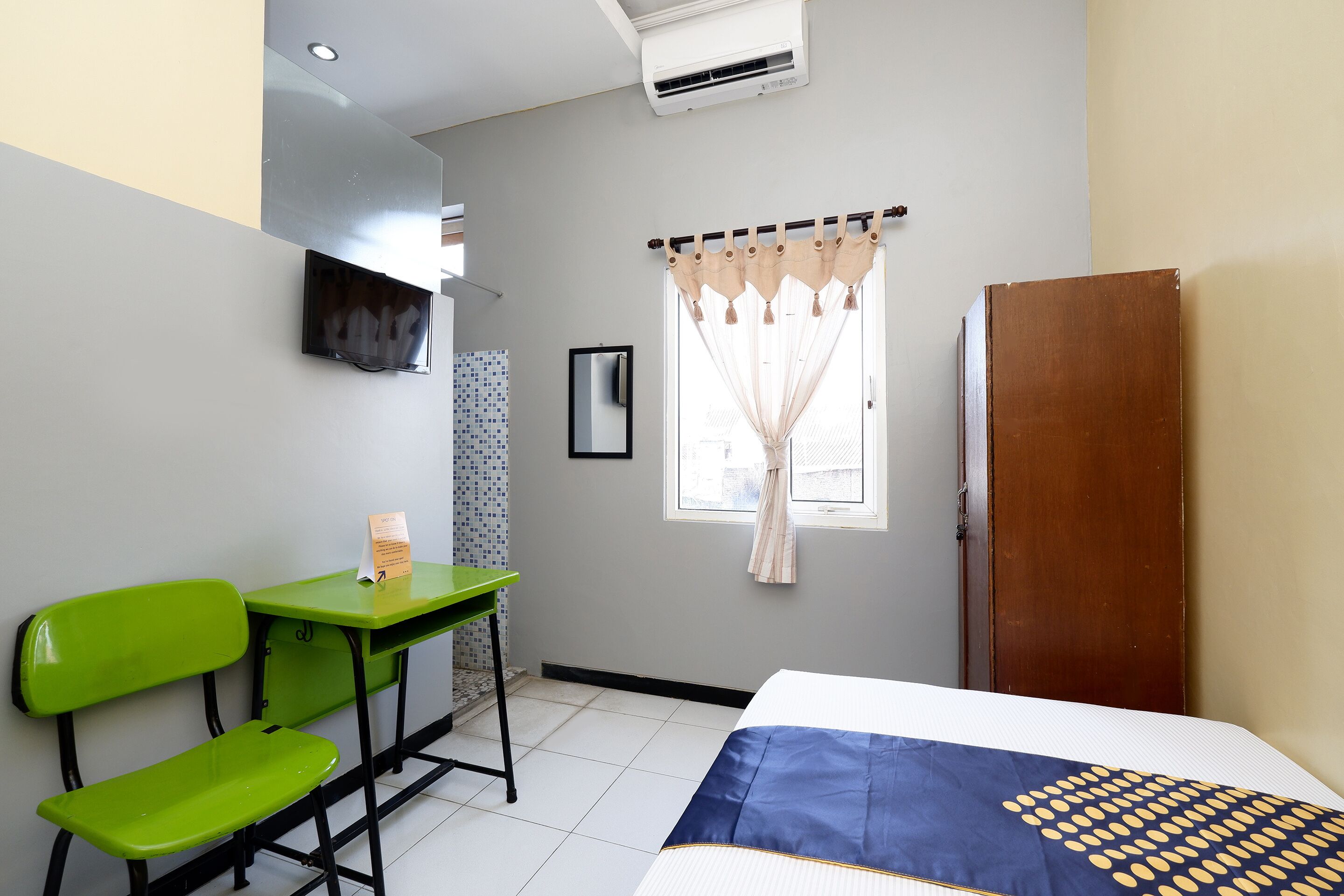 A room with a bed, a desk, two green chairs, a TV mounted on the wall, a window with curtains, and a wooden door.