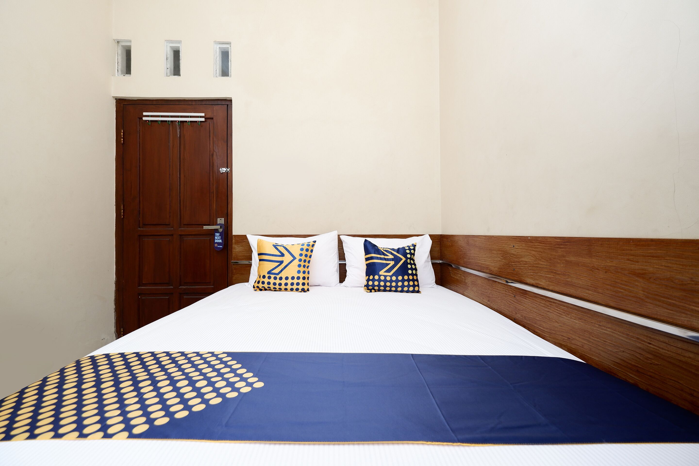 A hotel room with a wooden door, two pillows with geometric patterns, a blue and yellow bedspread, and a white bed.