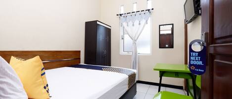 A hotel room with a bed, a green chair, a desk, a wardrobe, and a sign on the door.