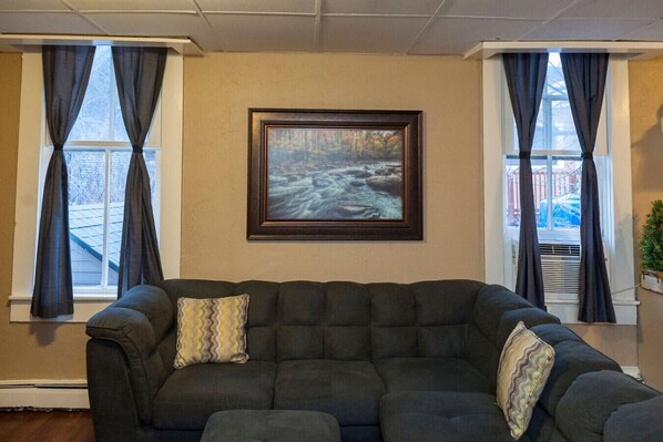 A living room with a dark-colored sofa, two windows with dark curtains, and a framed painting on the wall.