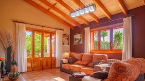 Ein gemütliches Wohnzimmer mit Holzbalkendecke, großen Fenstern mit Vorhängen, einer Couch und einem Tisch mit einer Vase.