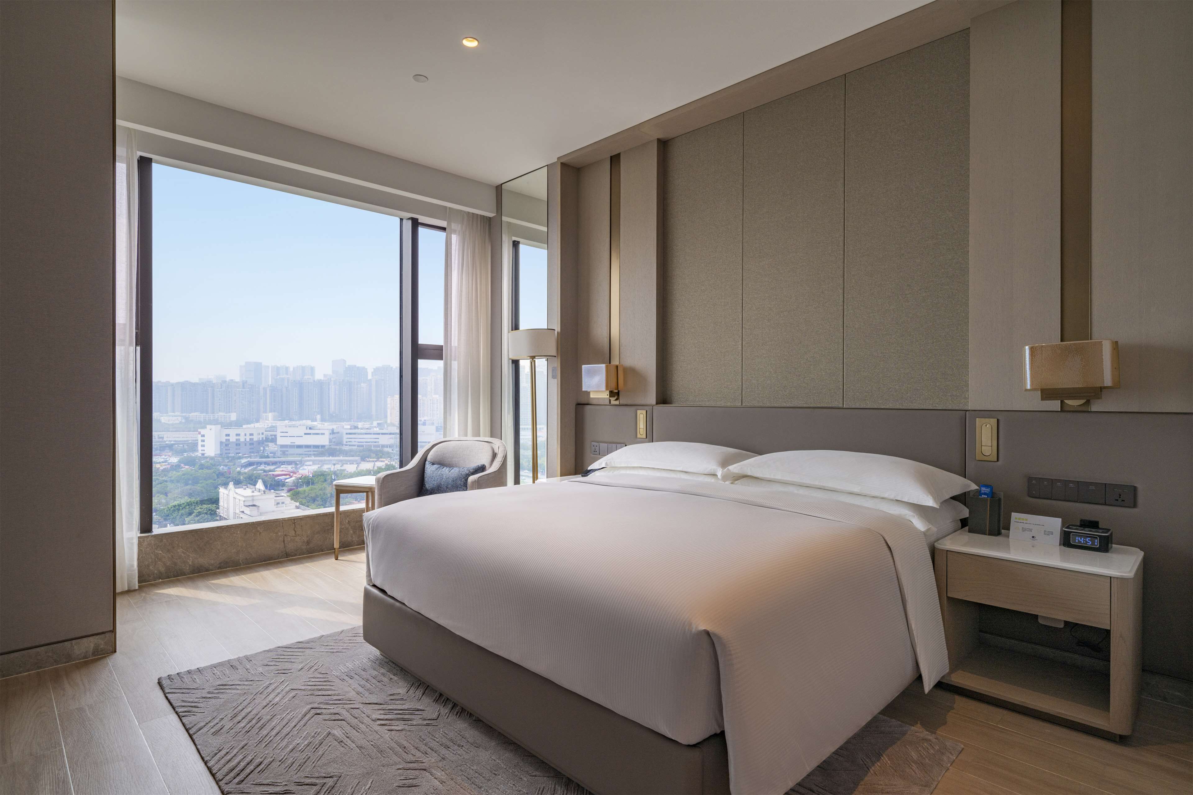 A modern hotel room with a large bed, a nightstand, a chair, and a view of the city through large windows.