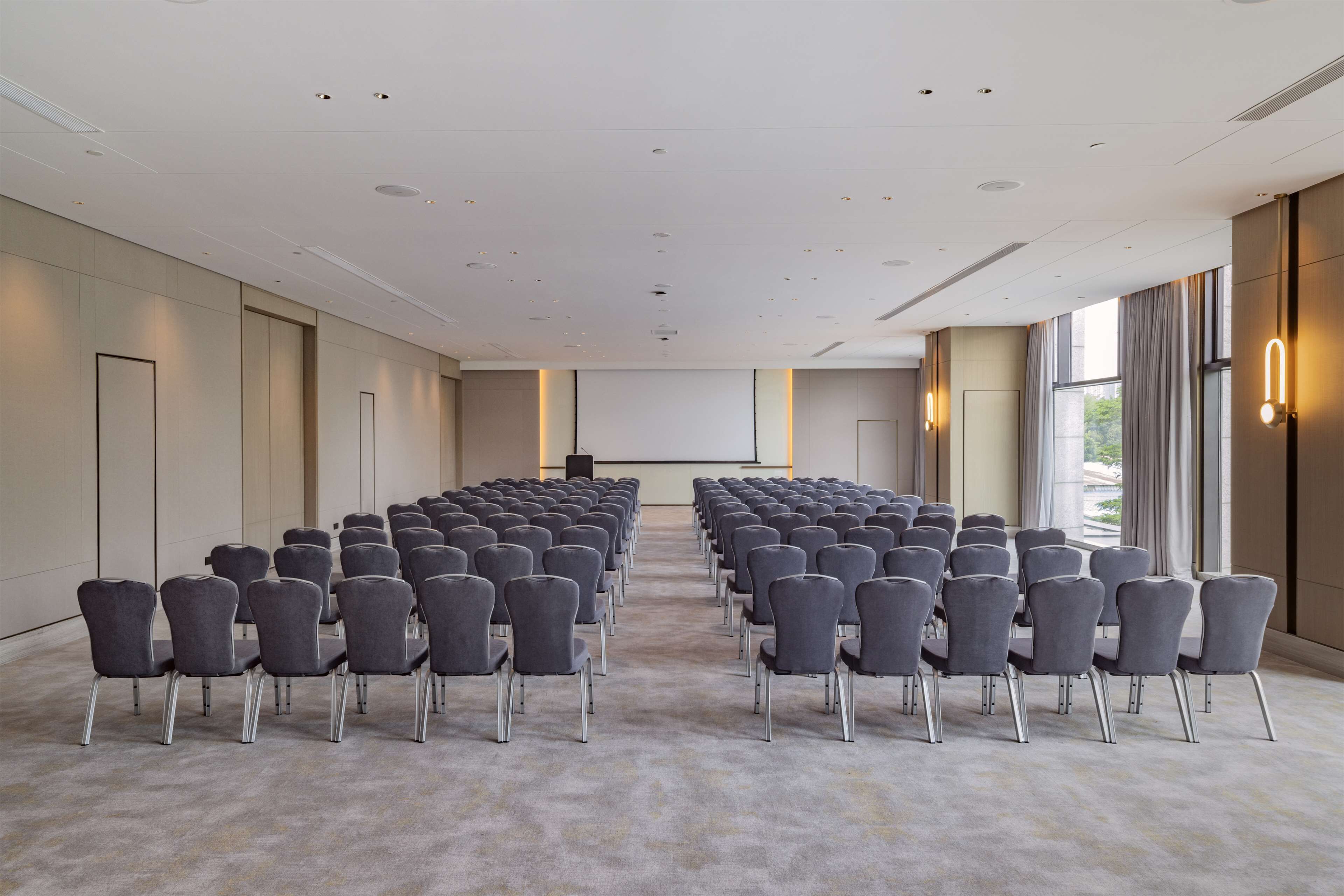Une salle de conférence avec des rangées de chaises disposées face à un écran de projection, de grandes fenêtres et une décoration aux couleurs neutres.