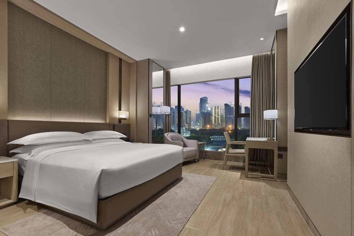 Ein modernes Hotelzimmer mit einem großen Bett, einem Schreibtisch und einem Sessel. Ein großes Fenster bietet Blick auf die Stadt.
