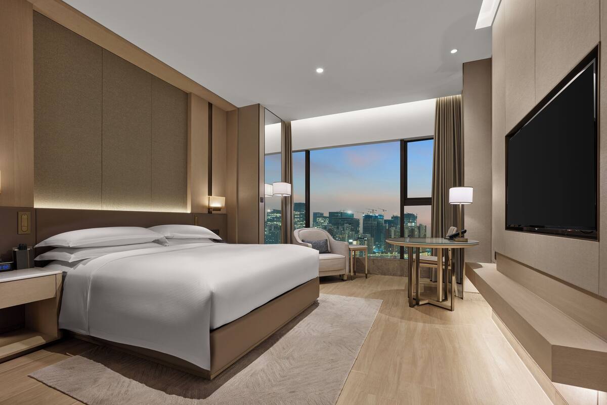 Ein modernes Hotelzimmer mit einem großen Bett, Nachttischen, einem Schreibtisch, einem Sessel sowie Blick auf die Stadt.