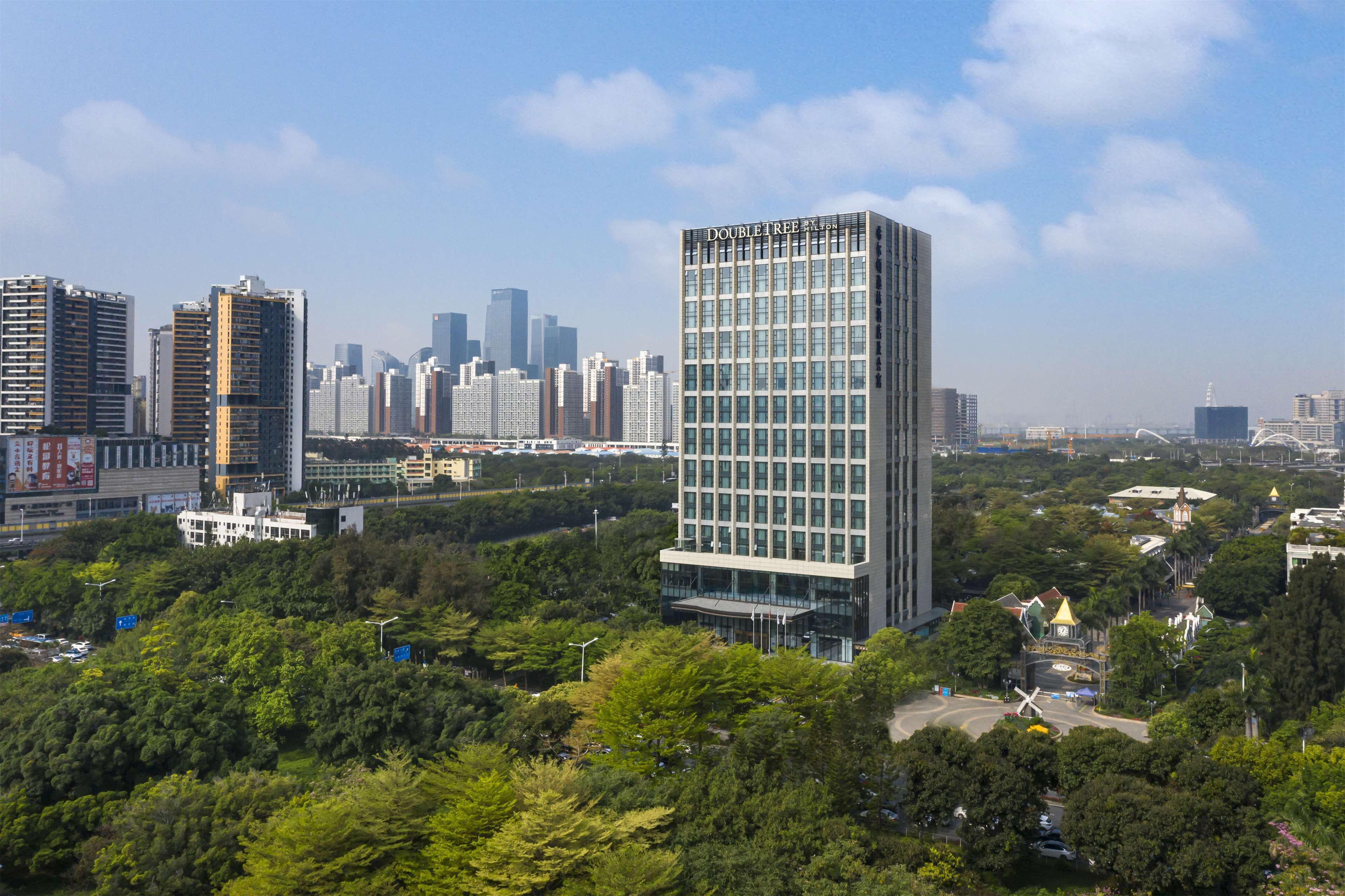 A modern high-rise building with a glass facade, surrounded by lush greenery and other high-rise buildings in the background.