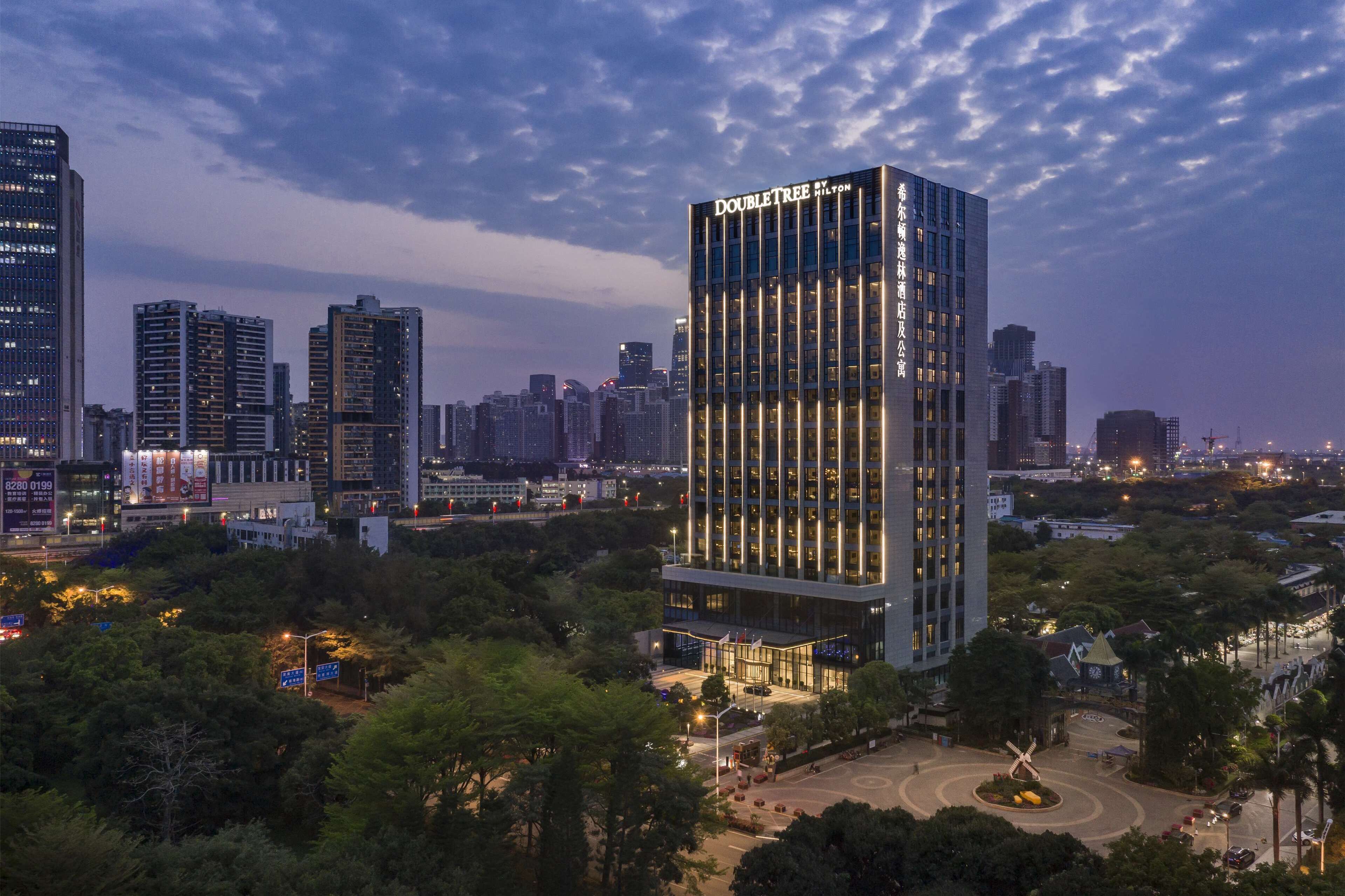 A modern hotel with a prominent "DoubleTree" sign, surrounded by lush greenery and other high-rise buildings in the background.