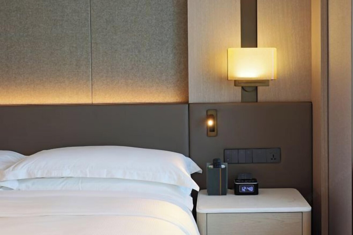 A neatly made bed with white linens, a bedside table with a lamp, and a digital clock displaying 12:48.