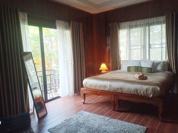 A bedroom with a large bed, wooden flooring, a nightstand with a lamp, and a view of the outdoors through the windows. - Baanbua Nonlen (Udon Thani)