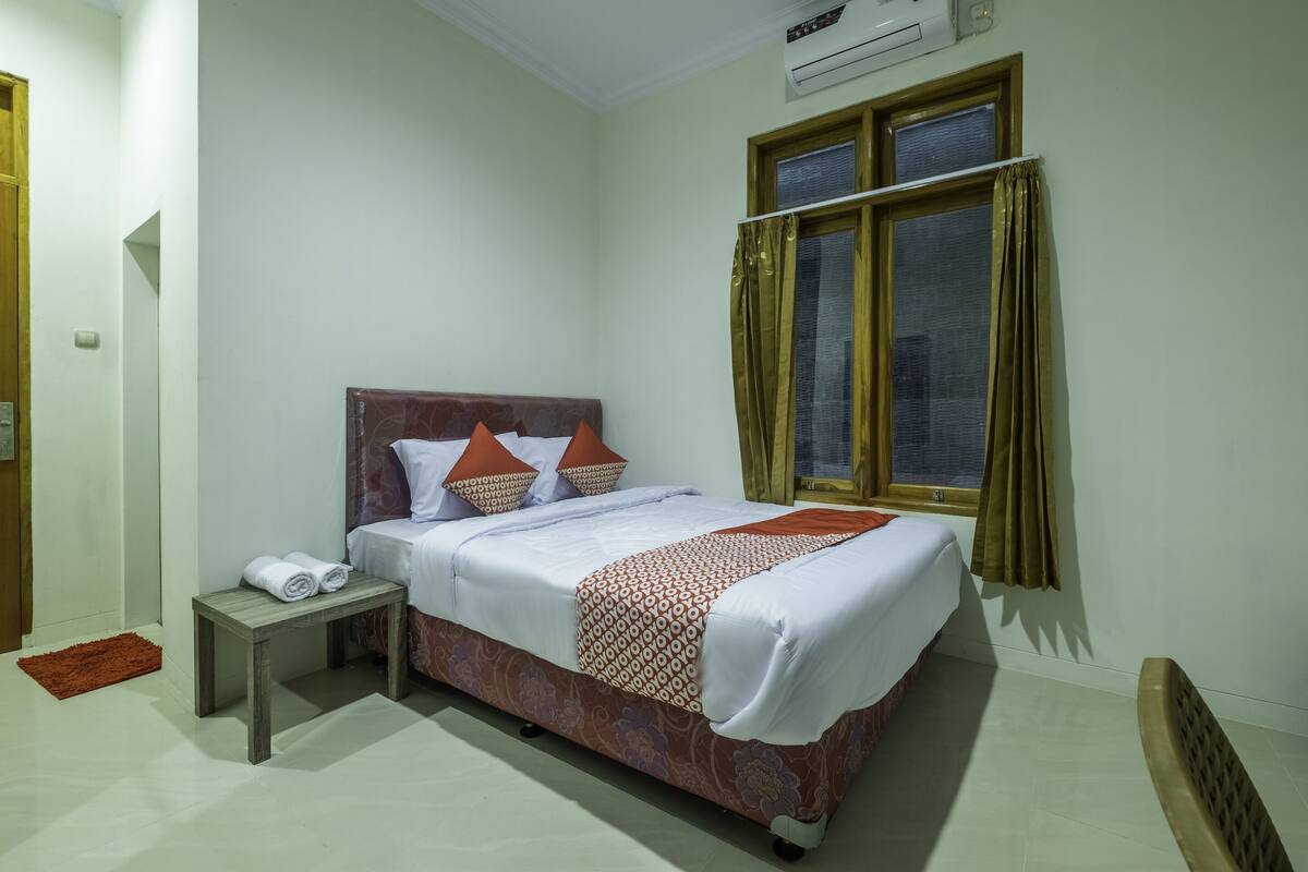 A neatly arranged hotel room with a bed, a bedside table with towels, a window with curtains, and an air conditioning unit.
