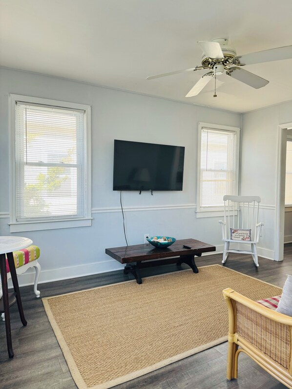 A living room with a flat-screen TV mounted on the wall, a wooden coffee table, a wicker chair, and a ceiling fan.