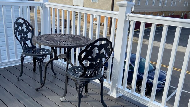 A wooden deck with a round table and two chairs, white railings, and a view of a parking lot and buildings in the background. - Beach Block, Renovated 2 bdrm, sleep 6, 2nd Flr unit Parking and 4 beach badges (Seaside Park)