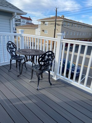 A wooden deck with a round table and two chairs, white railings, and a view of a parking lot and buildings in the background.
