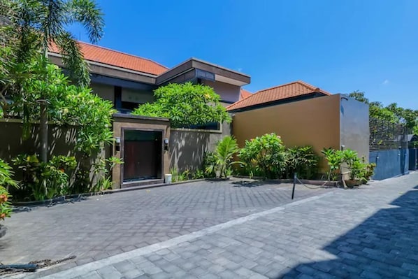 Una zona residencial con camino de entrada pavimentado, una casa con tejado de tejas rojas y vegetación circundante.