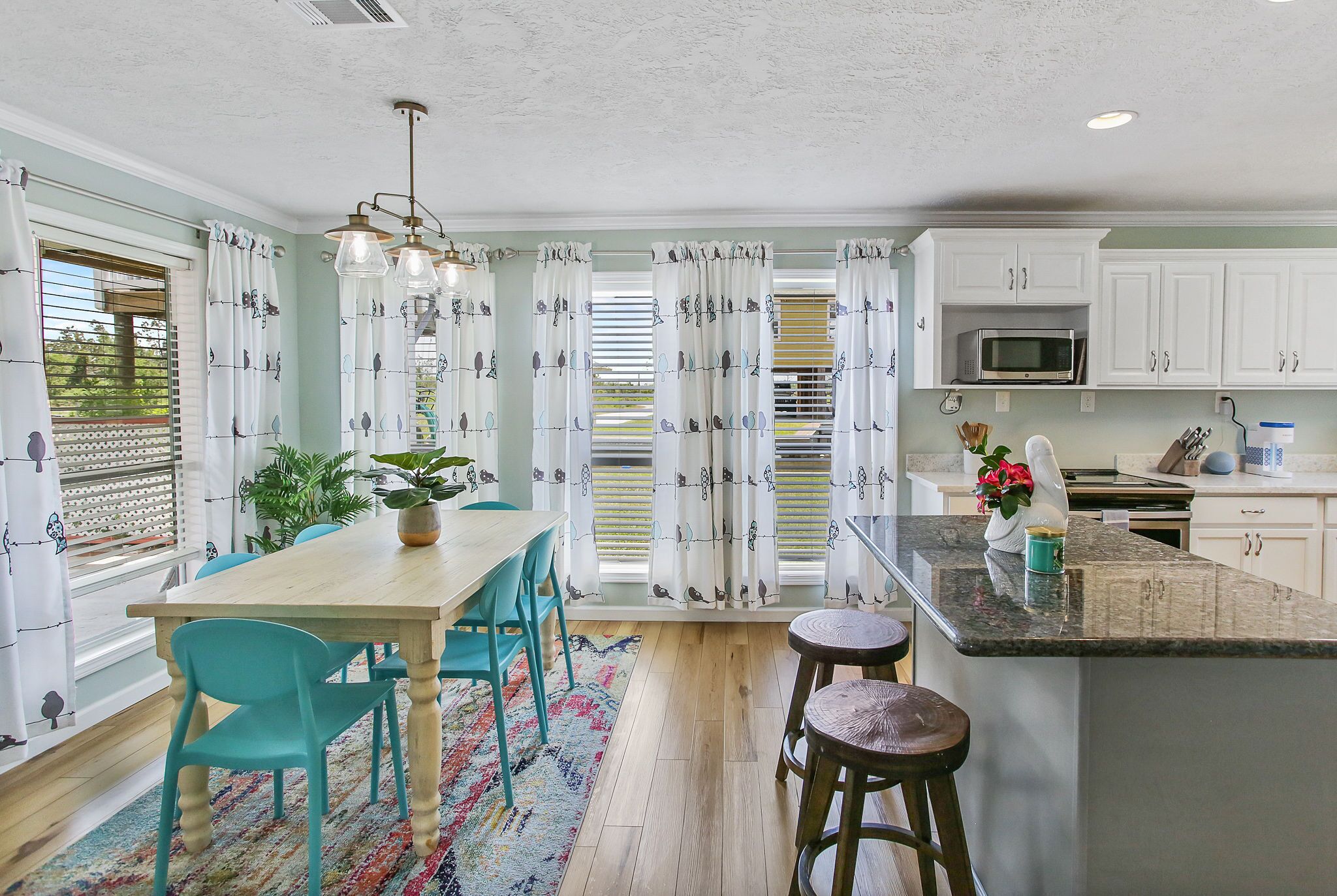 A kitchen with a wooden table, teal chairs, a colorful rug, and white cabinets.