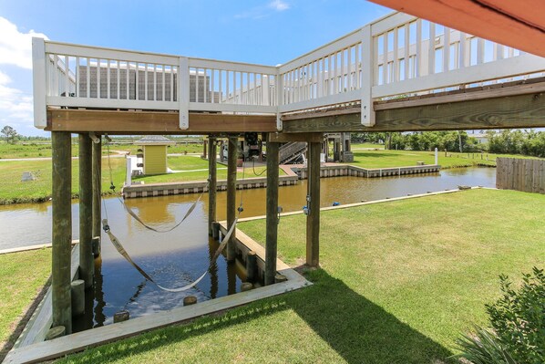 A deck structure with a wooden floor and railings, supported by wooden posts, overlooking a water feature and a grassy area.