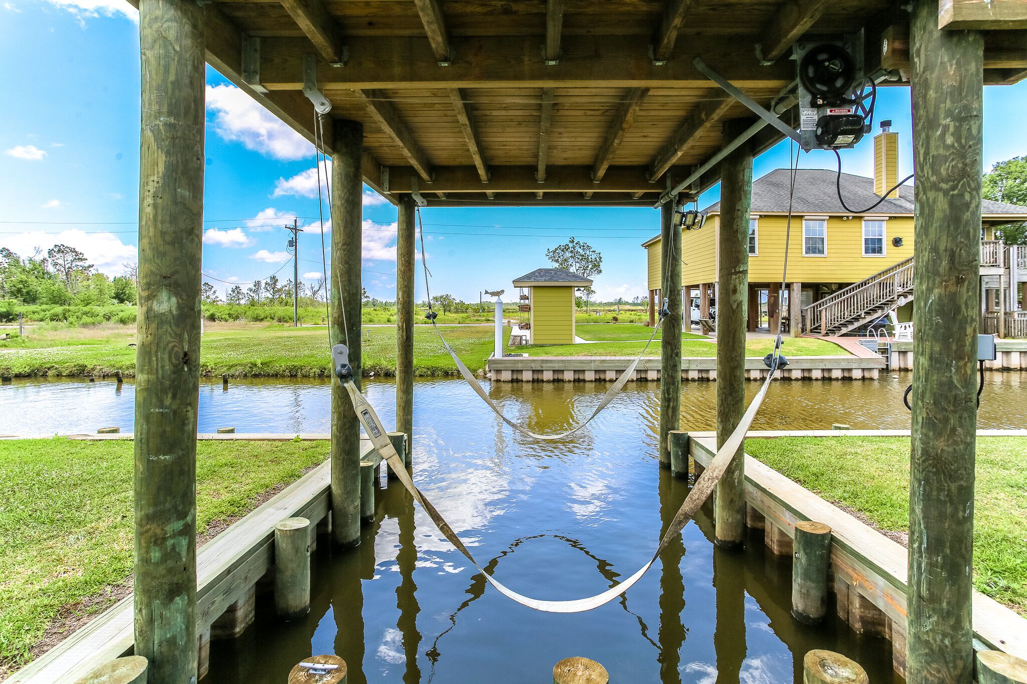 A wooden structure with a hammock over a water canal, a small shed, and a yellow house in the background.