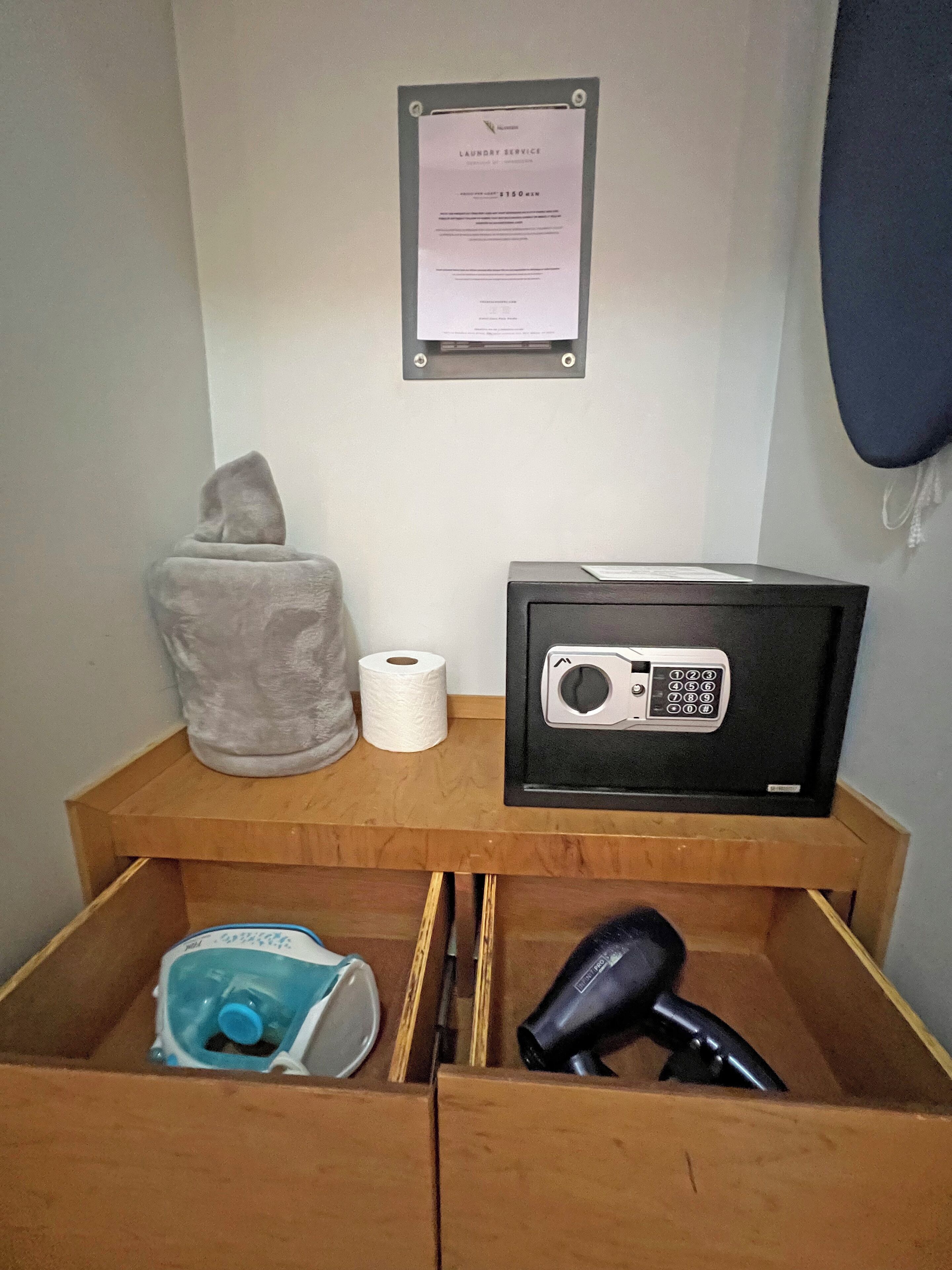 A small wooden cabinet with a framed notice, a gray towel, a roll of toilet paper, and a black electronic device.