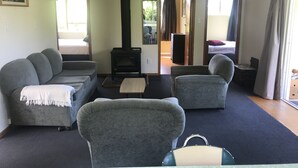 En stue med sofa, lænestole, sofabord og pejs. Der er en seng i baggrunden og et vindue med gardiner.