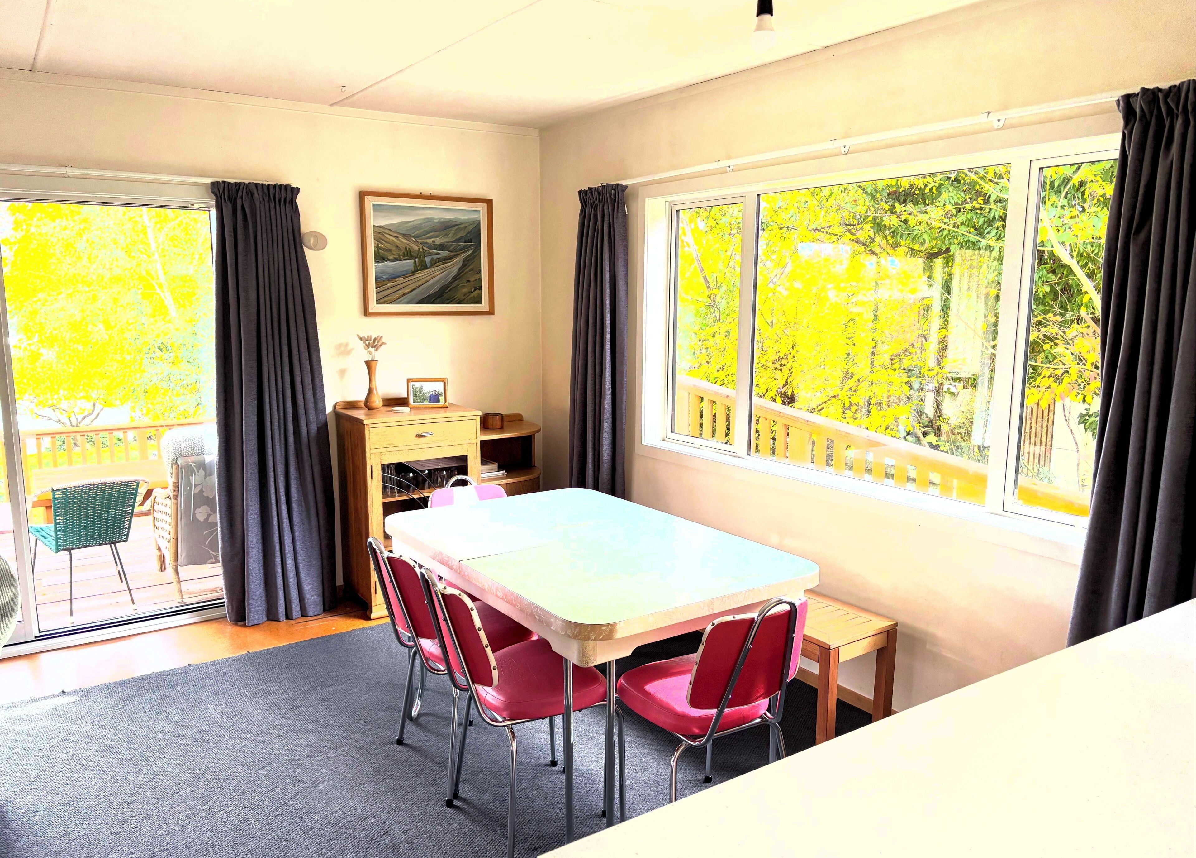 A room with a dining table, chairs, a small desk, a chair, a window with curtains, and a painting on the wall.