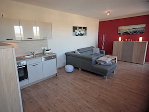 Une cuisine moderne avec des meubles blancs, un lave-vaisselle et une cuisinière. On y trouve un canapé gris avec des coussins et une petite table sur laquelle est posée une bougie. Un mur rouge orné d’un tableau est visible.
