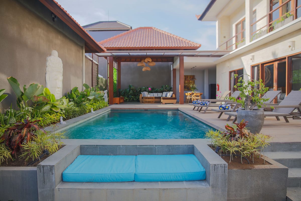 A swimming pool with a blue cushioned seating area, surrounded by plants and lounge chairs.