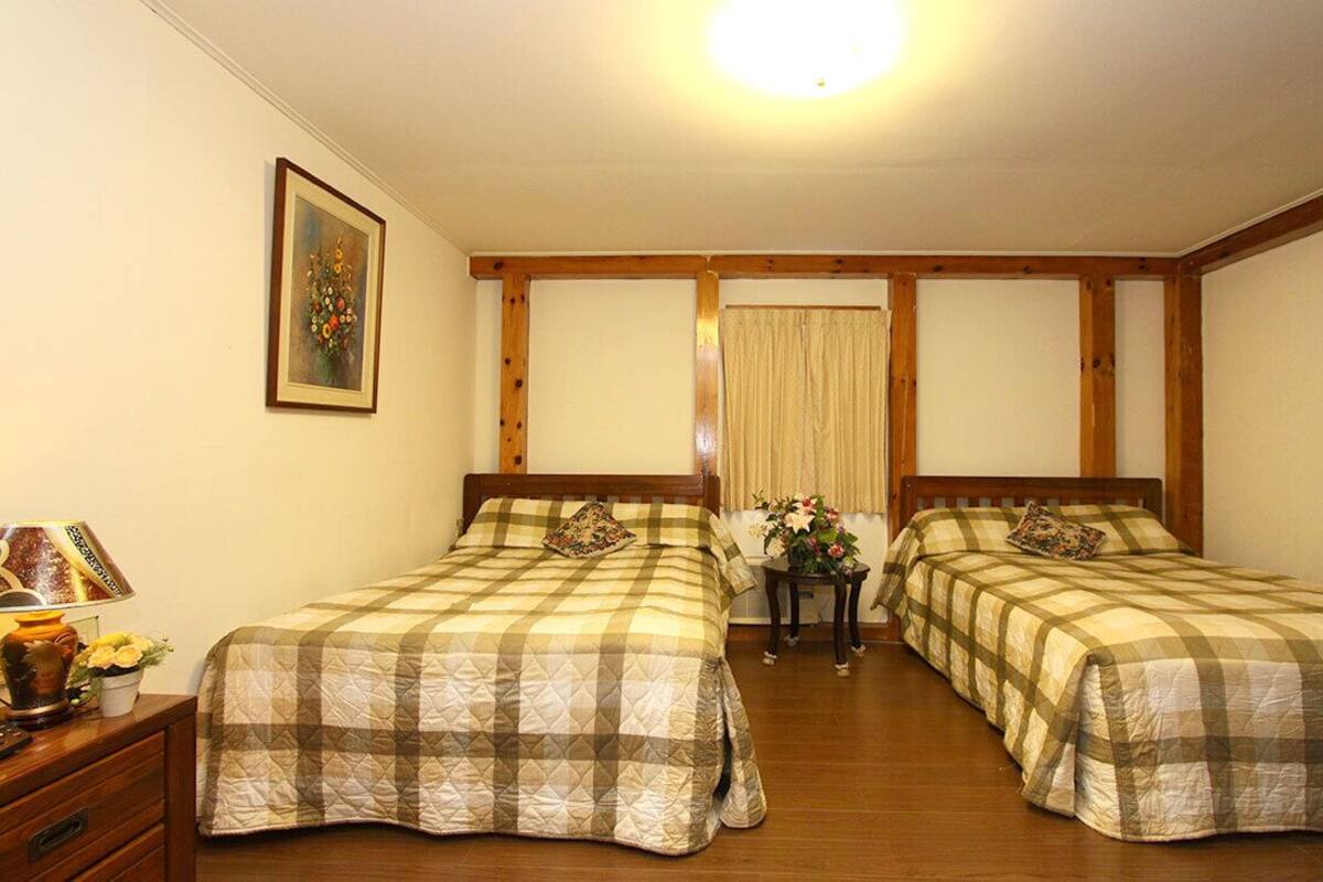 A room with two beds, wooden beams, a painting, a lamp, and a table with flowers.