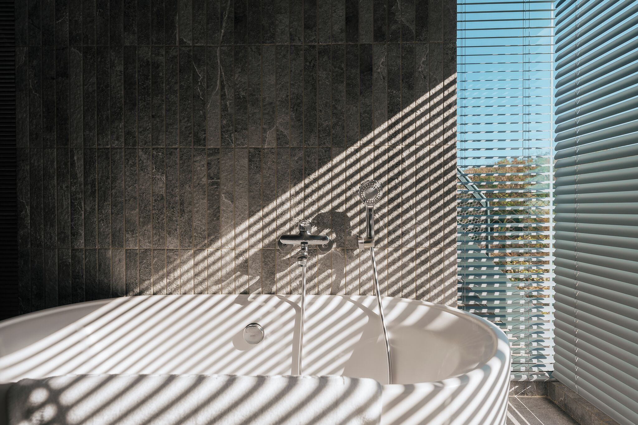 A modern bathroom with a freestanding bathtub, a shower with a handheld showerhead, and a window with blinds.