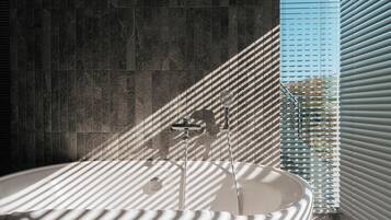 A modern bathroom with a freestanding bathtub, a shower with a handheld showerhead, and a window with blinds.