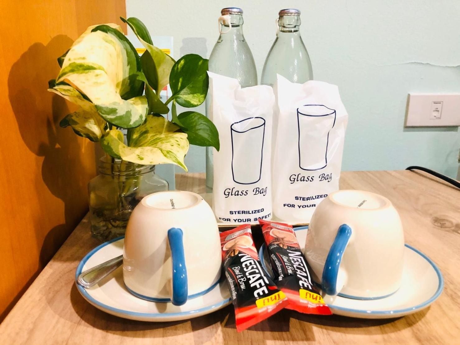 Un tavolo con due tazze bianche con manici blu, un vaso con foglie verdi, due bottiglie di vetro e due sacchetti con l'etichetta 'Glass Bag' e la scritta 'STERILIZZATO PER LA TUA SICUREZZA'.