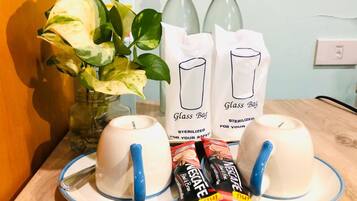 Un tavolo con due tazze bianche con manici blu, un vaso con foglie verdi, due bottiglie di vetro e due sacchetti con l'etichetta 'Glass Bag' e la scritta 'STERILIZZATO PER LA TUA SICUREZZA'.