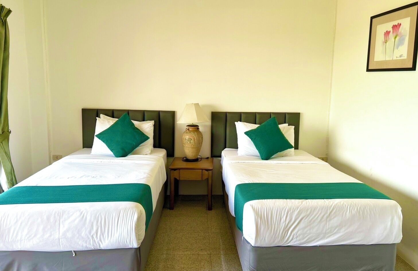 A hotel room with two single beds, each with white linens and green throw pillows. There is a nightstand between the beds with a lamp and a vase. The room has a simple, clean design with a light-colored wall and a framed picture above the bed.