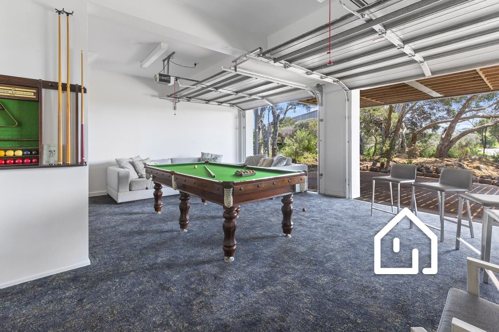 A pool table in a room with a blue carpet, white walls, and a view of trees and sky.