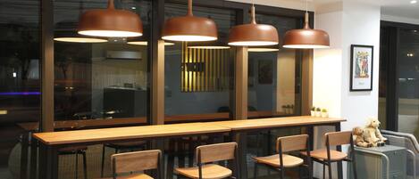 Un área de bar moderna con mesas y sillas de madera, iluminación colgante y amplios ventanales.