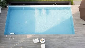 Una piscina rectangular con un muelle de madera, una silla de descanso blanca y una pequeña mesa con pelotas de colores.