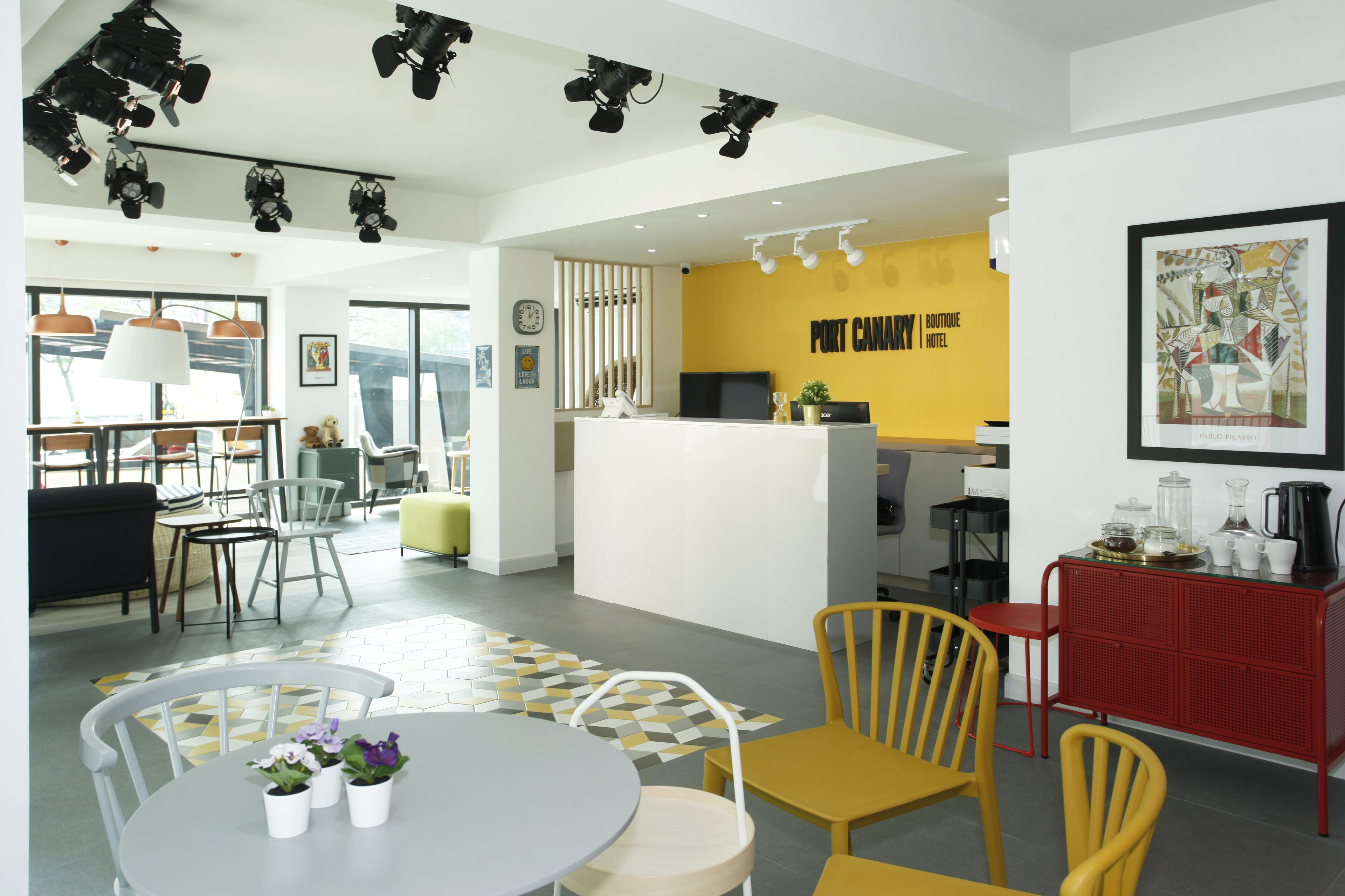 Un hall d’hôtel moderne avec un mur d’accent jaune, un comptoir d’accueil et un espace repas doté d’un sol à motifs géométriques.