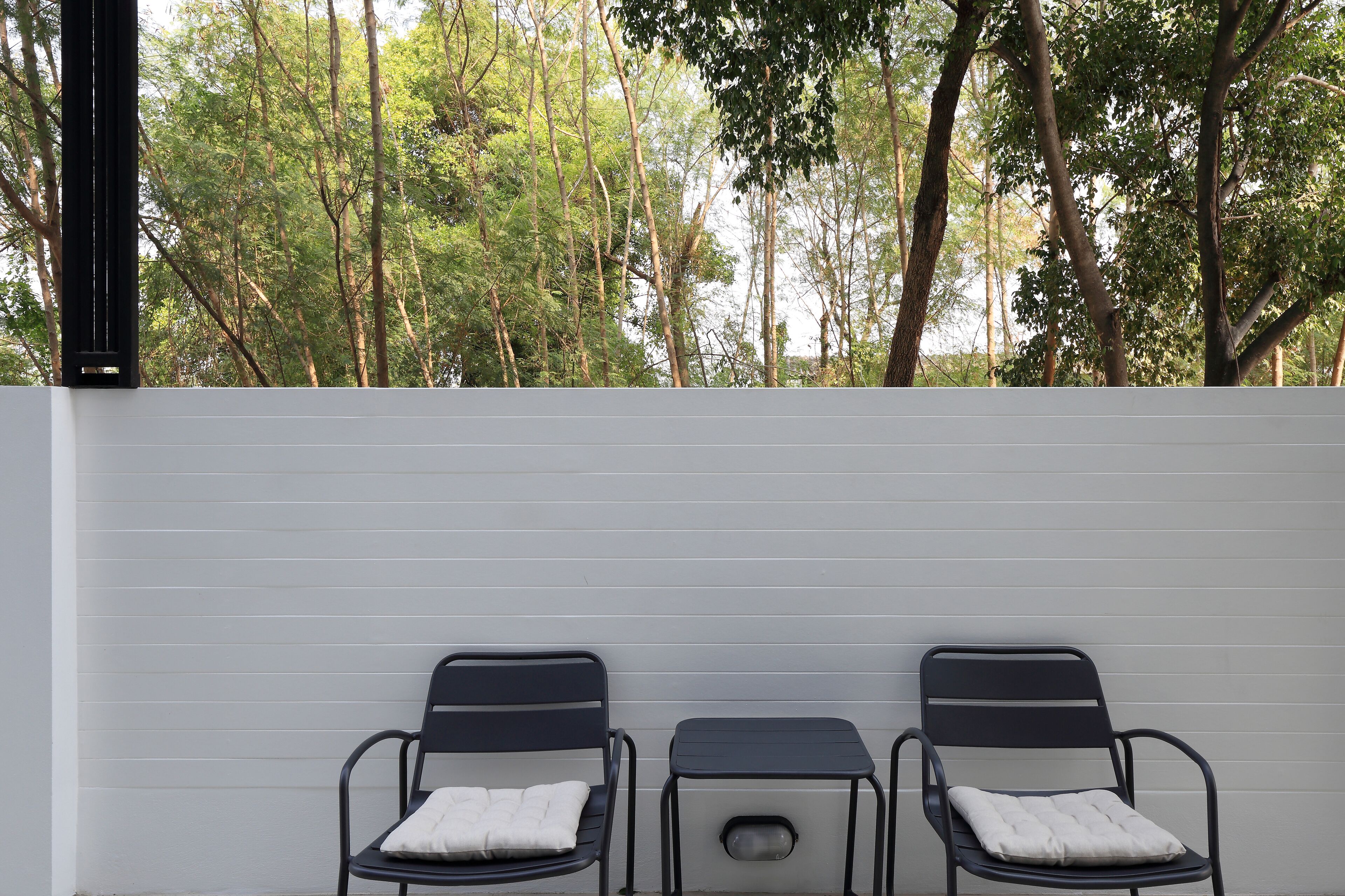 Two black metal chairs with white cushions against a white wall, with a view of trees in the background.