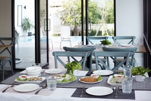 A dining table set with plates, bowls, and glasses, with a view of an outdoor area including a pool and chairs. - Port Canary Hotel (Bangkok)