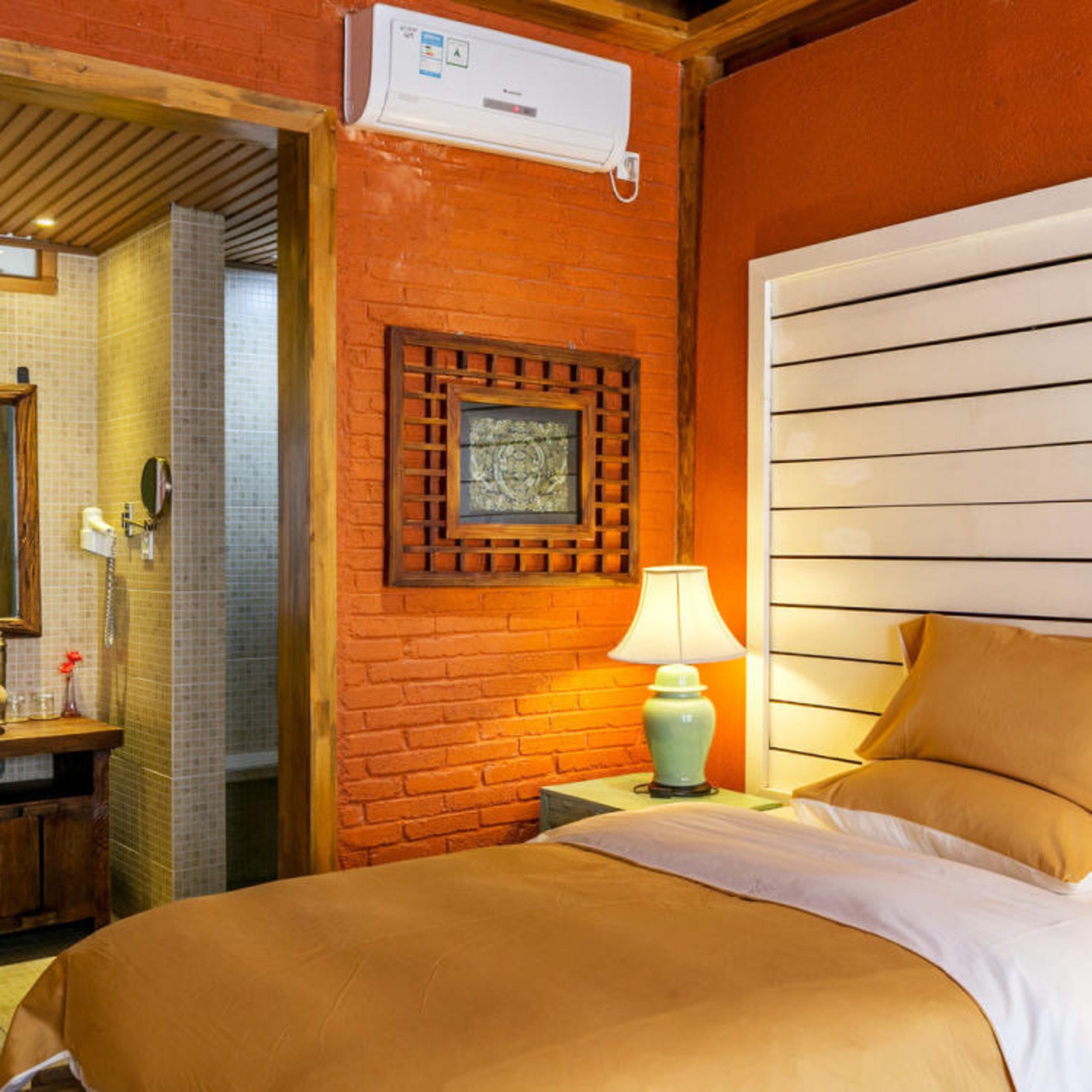 A hotel room with a brick wall, a bed with white bedding, a nightstand with a lamp, and a window with horizontal blinds.