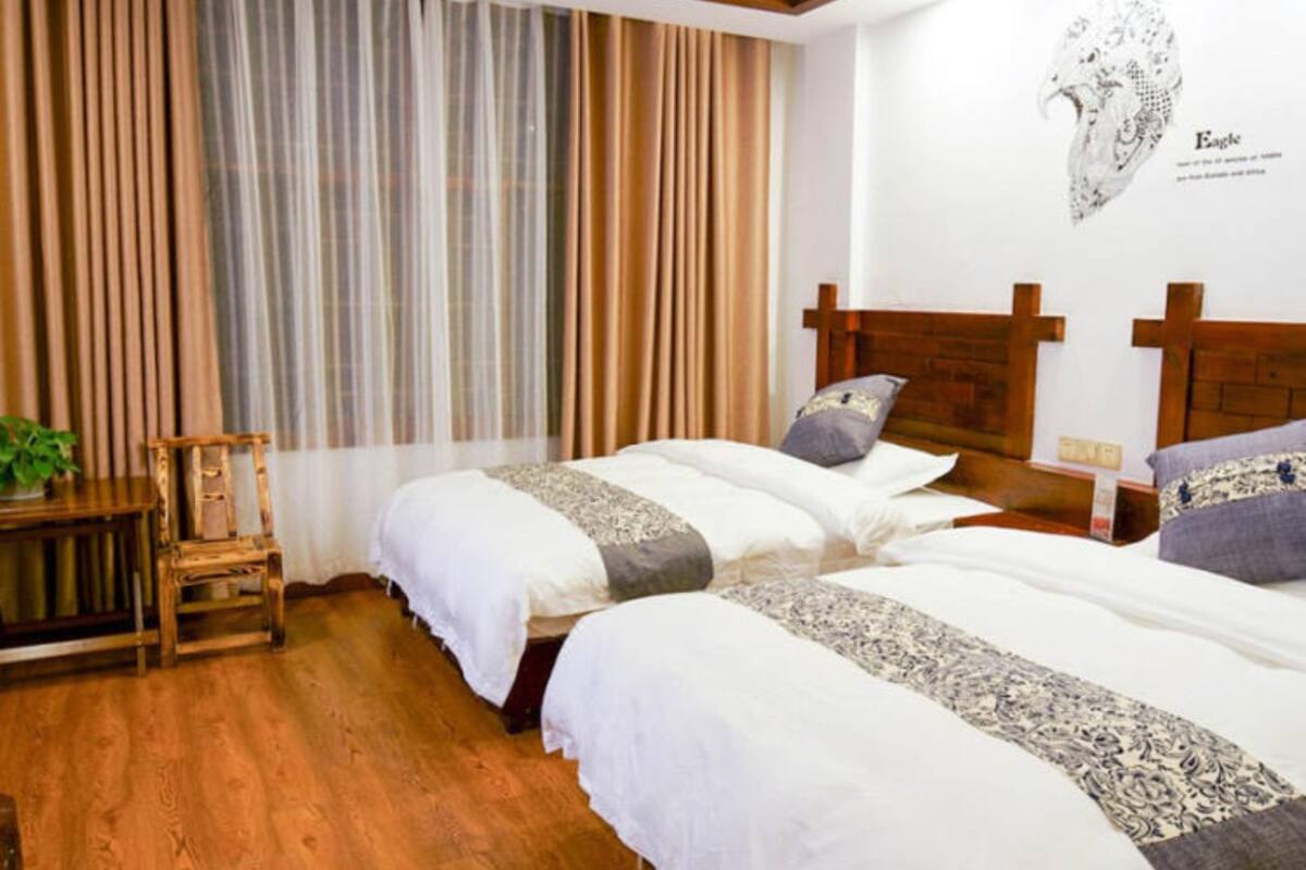 A room with two beds, wooden flooring, a window with curtains, and a wall-mounted artwork.