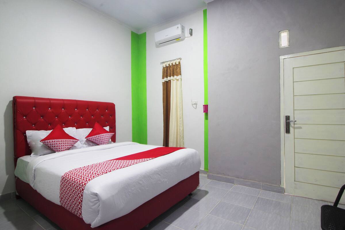 A hotel room with a red upholstered bed, white bedding, a green accent wall, a white door, and a window with curtains.