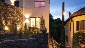 A modern building with a stone facade, a wooden fence, and a gravel path leading to a seating area with chairs and a table. - Retreat in Malcantone (Alto Malcantone)