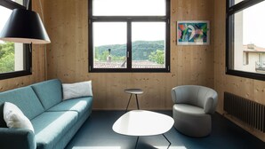 A modern interior with a wooden ceiling, a large window with a view of greenery, a blue sofa, a white table, and a gray armchair. - Retreat in Malcantone (Alto Malcantone)