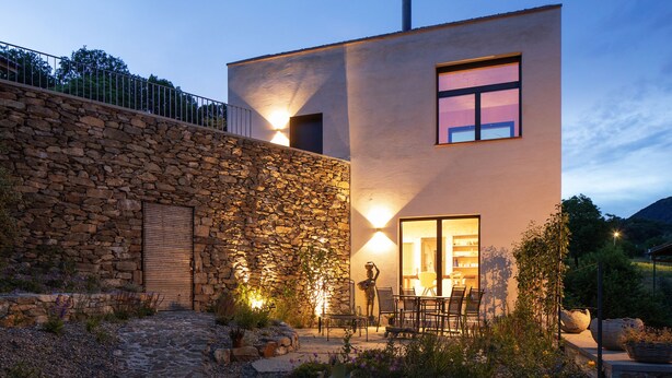 A modern house with a stone wall, a patio with chairs, and a view of the surrounding landscape. - Retreat in Malcantone (Alto Malcantone)