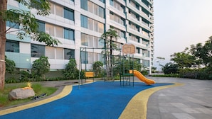 A playground with a blue and yellow play area, a basketball hoop, and a slide, surrounded by greenery and a modern building. - OYO 2245 Apartment Grand Kamala Lagoon (Bekasi)