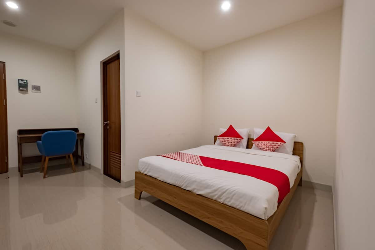 A bedroom with a wooden bed, white bedding, red accents, a desk, and a blue chair.