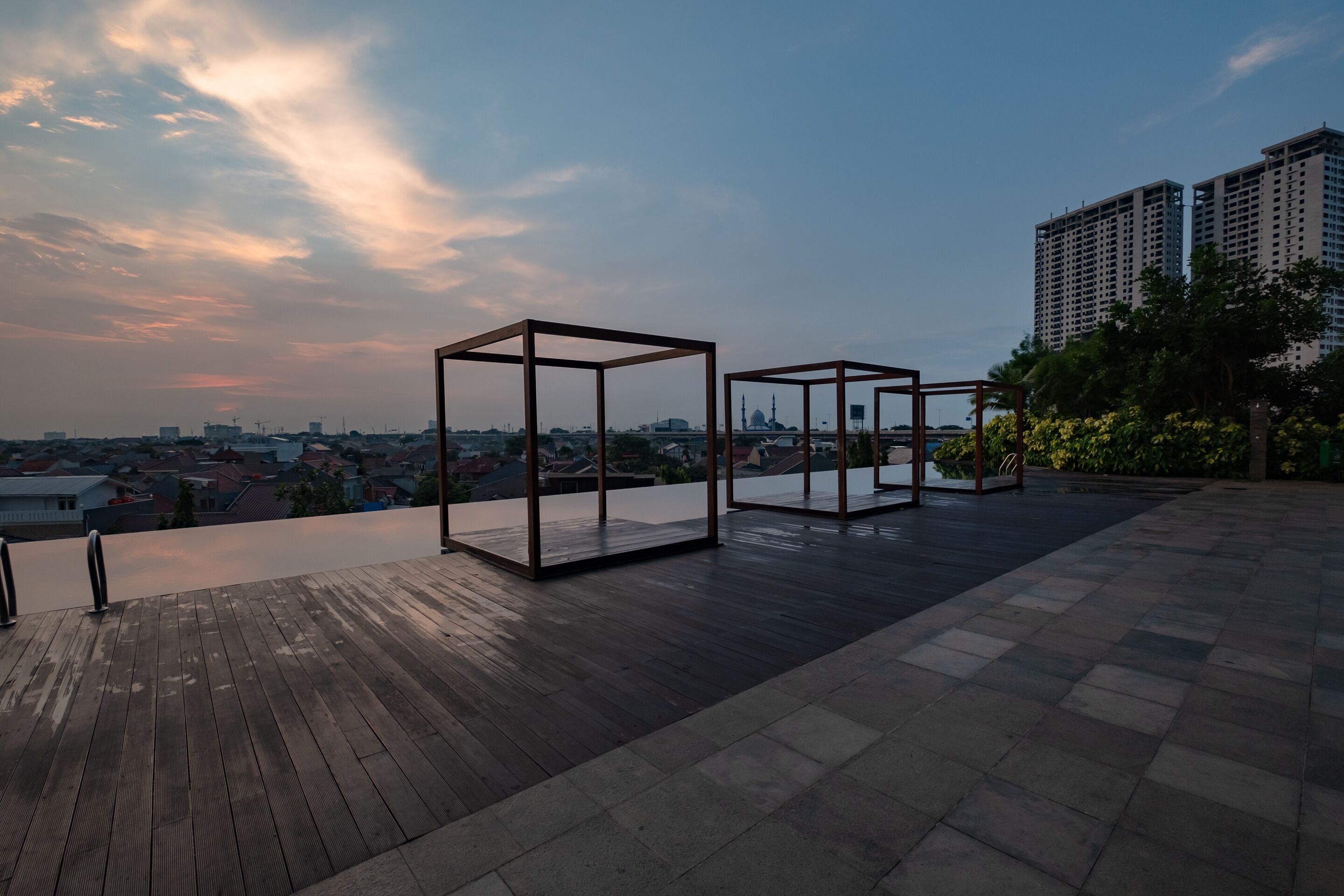 Une terrasse sur le toit, dotée d’un plancher en bois et de structures métalliques, offrant une vue sur un paysage urbain avec ses bâtiments et une mosquée.